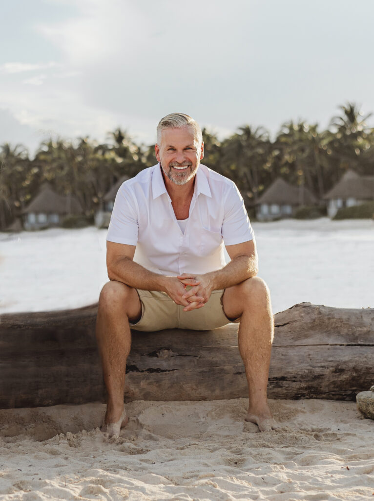 Man sitting on a log at the beach, smiling at the camera.